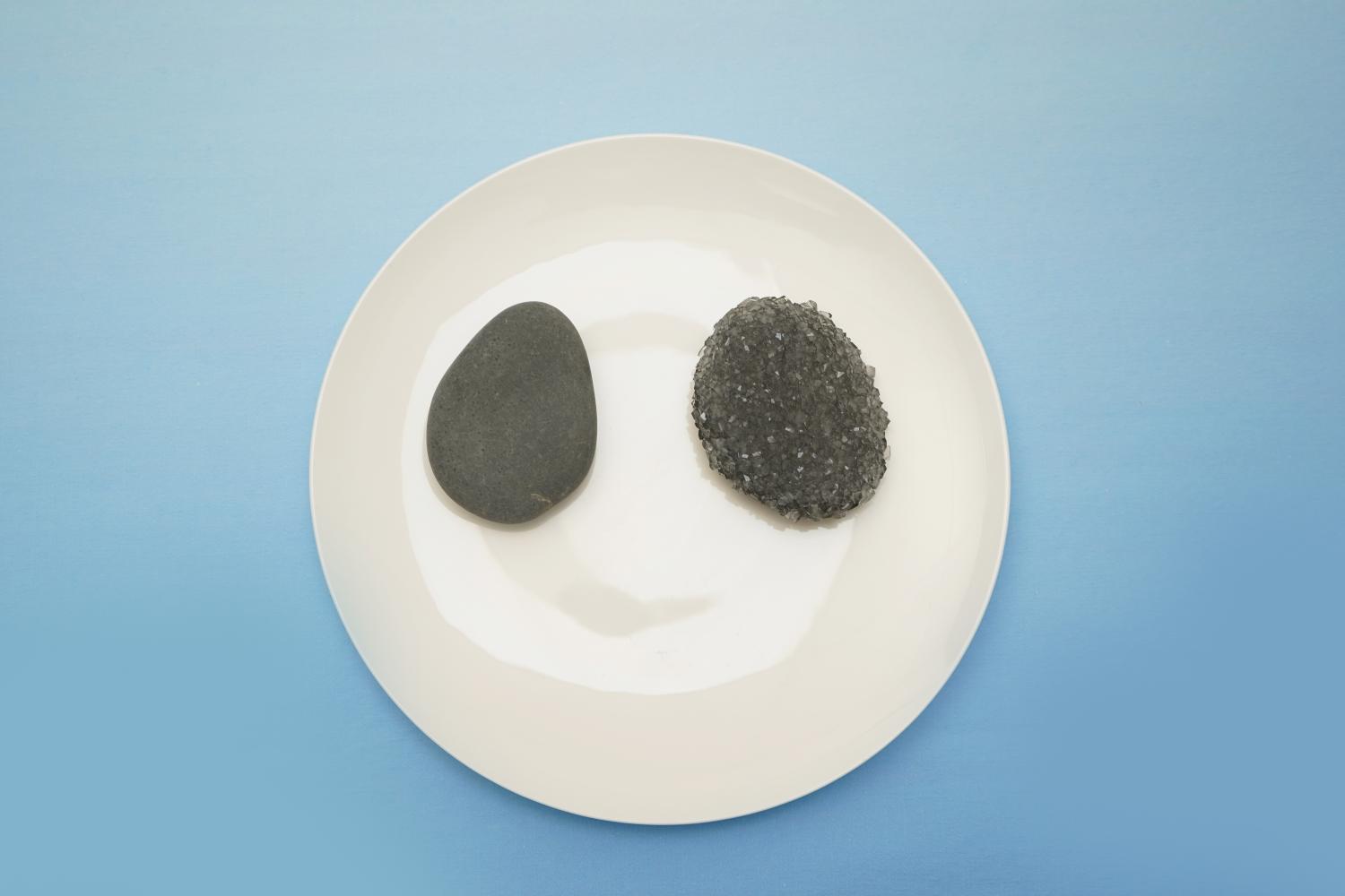 A smooth, grey stone and a cluster of grey crystals rest on a white plate against a pale blue background.