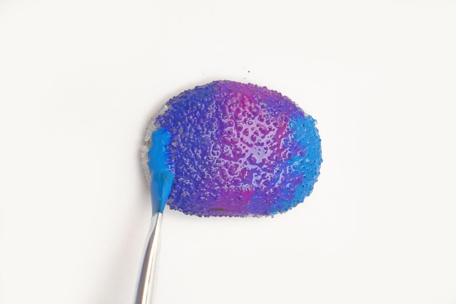 A paintbrush applies blue and purple paint to a textured, grey rock on a white surface.