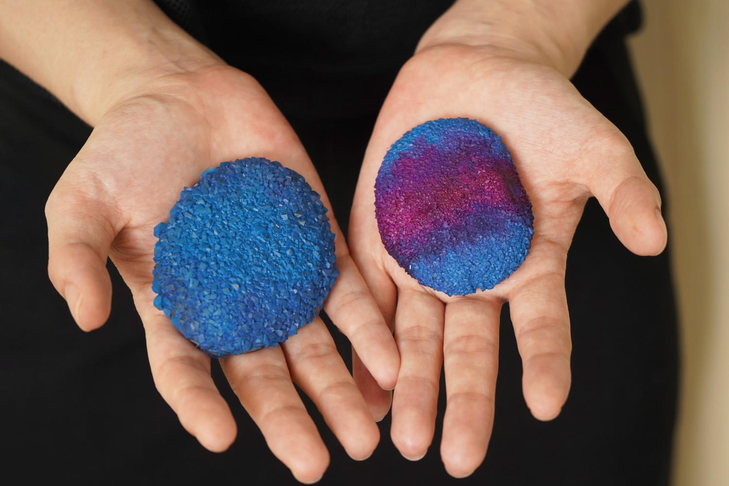 Two hands display painted rocks, one a solid blue, the other a blend of purple and blue, resembling crystals.