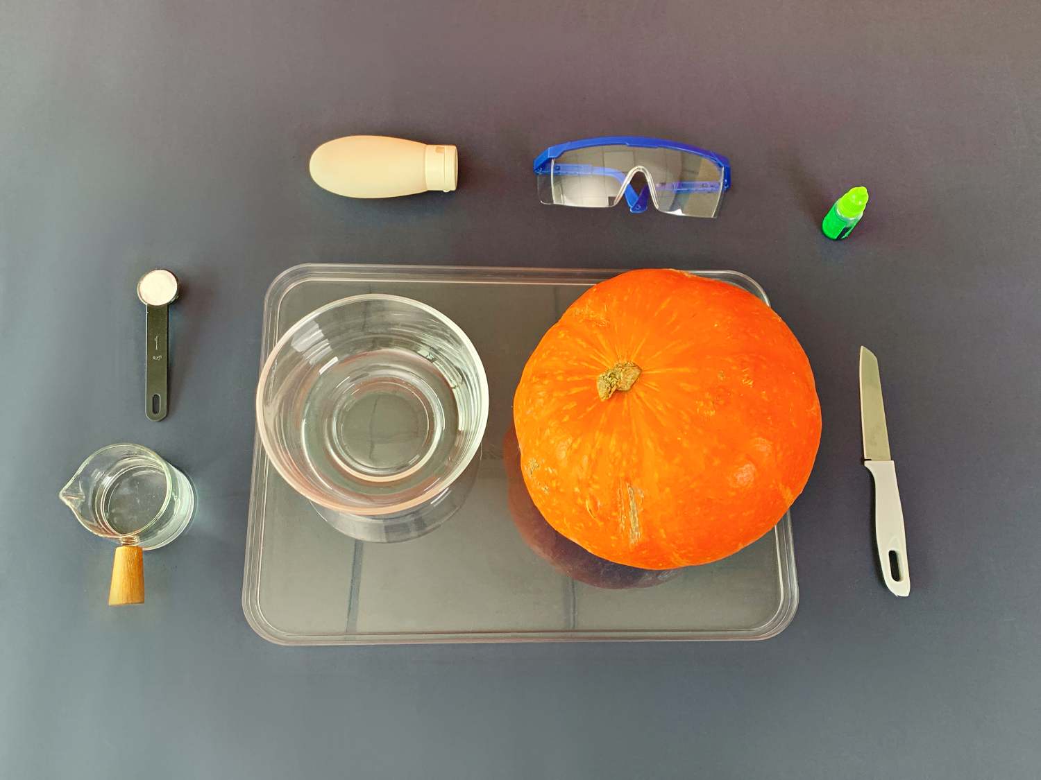 A whole pumpkin, a bowl of water, a small bottle of green liquid, safety goggles, a knife, a measuring spoon with white powder, and a small beaker of clear liquid are arranged on a clear tray placed on a dark gray surface. A sheet of green paper, a roll of tape, scissors, a pencil, a ruler, some gold string, and a pile of small, colourful pom-poms are arranged on a light wooden surface.