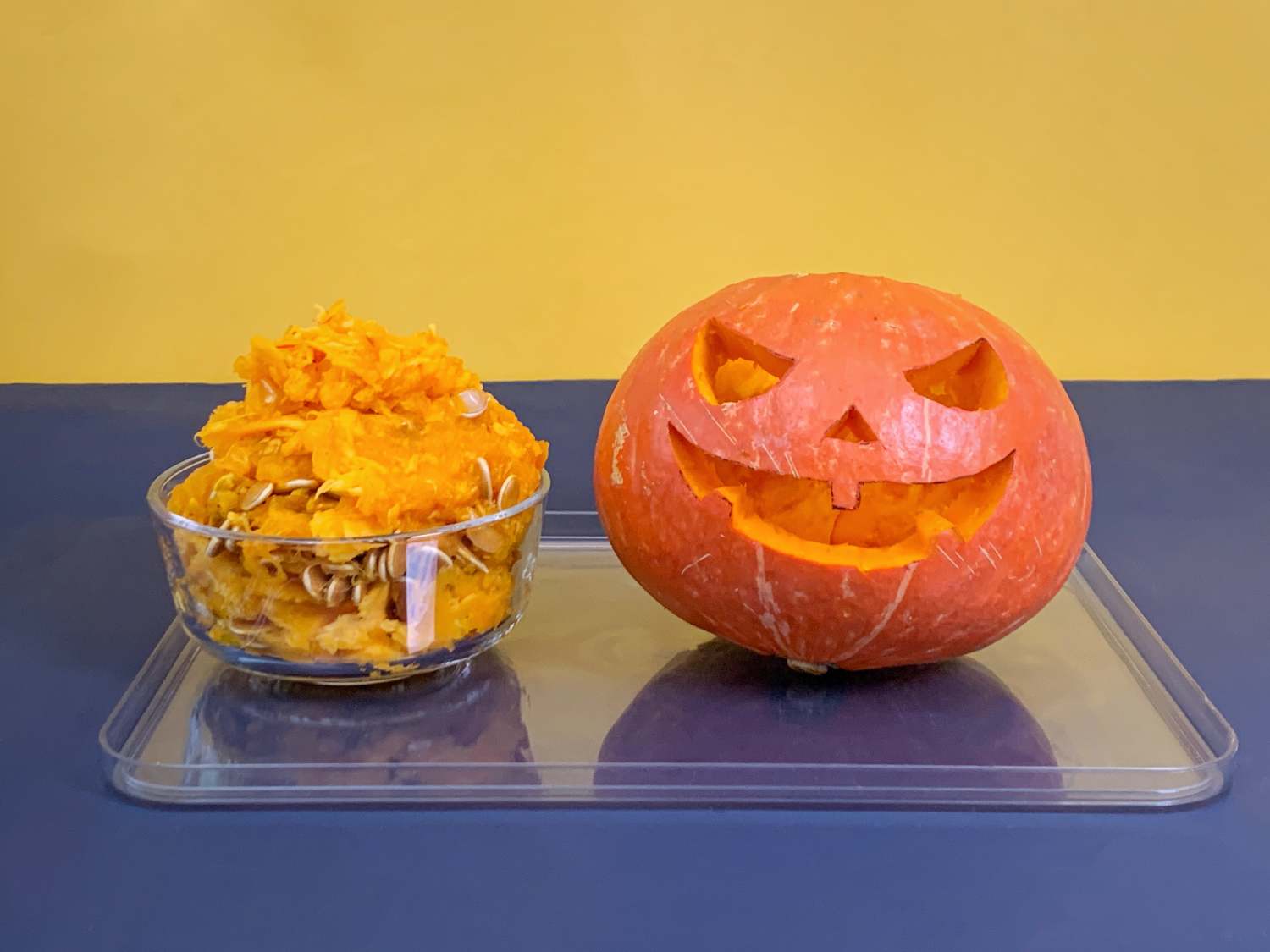 A carved pumpkin with a spooky face sits on a clear plastic tray next to a clear glass bowl of pumpkin pulp and seeds, against a two-toned yellow and dark blue background.