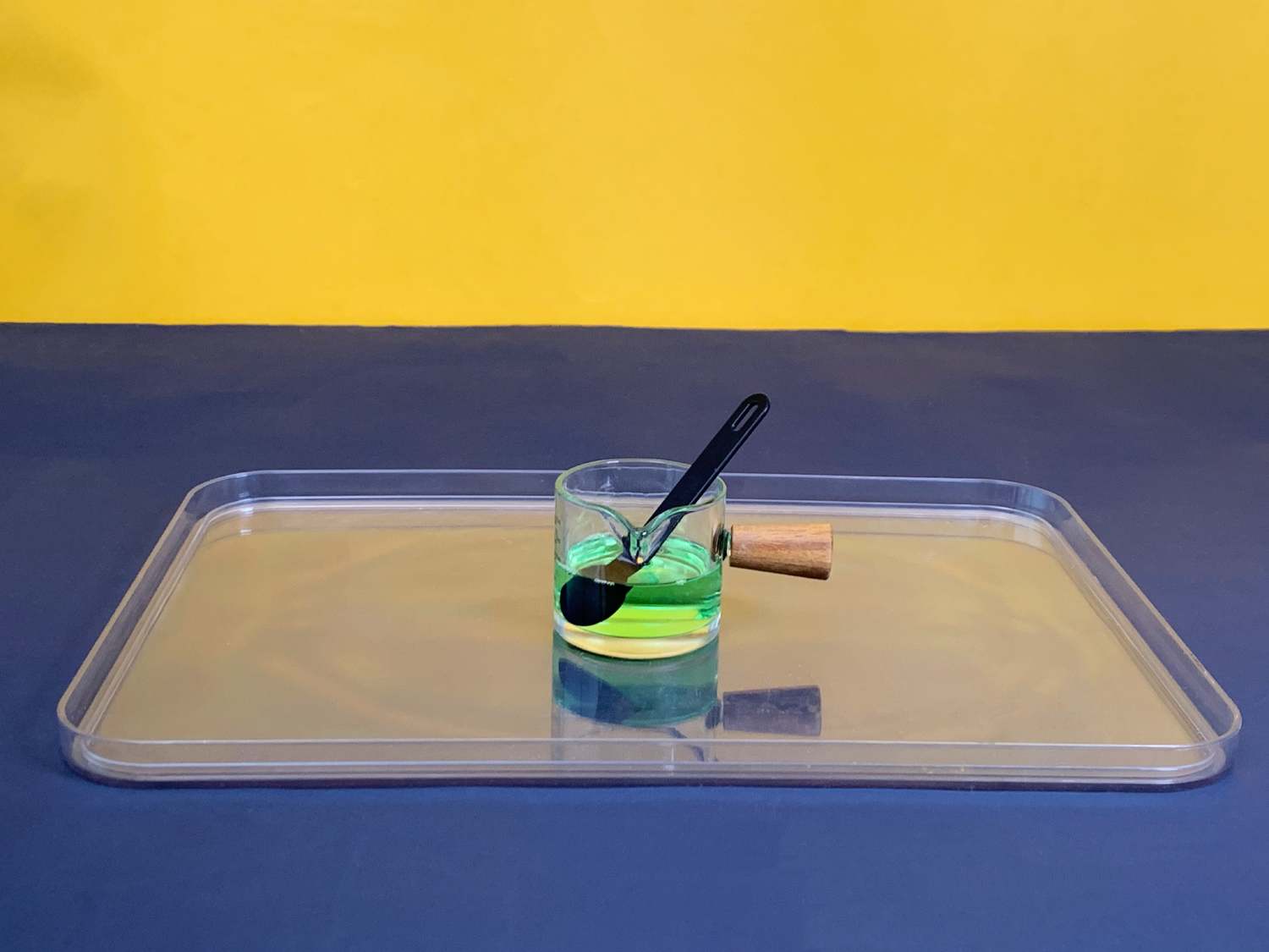 A small, clear glass container with a wooden handle, containing a bright green liquid and a black spoon, sits on a clear plastic tray against a two-toned yellow and dark blue background.