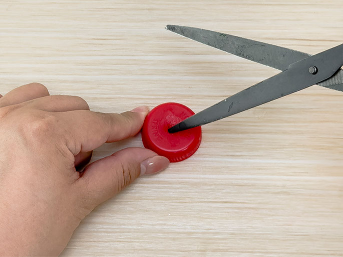 A hand holds a red plastic bottle cap on a light wooden surface while a pair of scissors pierces the center of the cap. The hand's fingers gently secure the cap, indicating a careful cutting process.