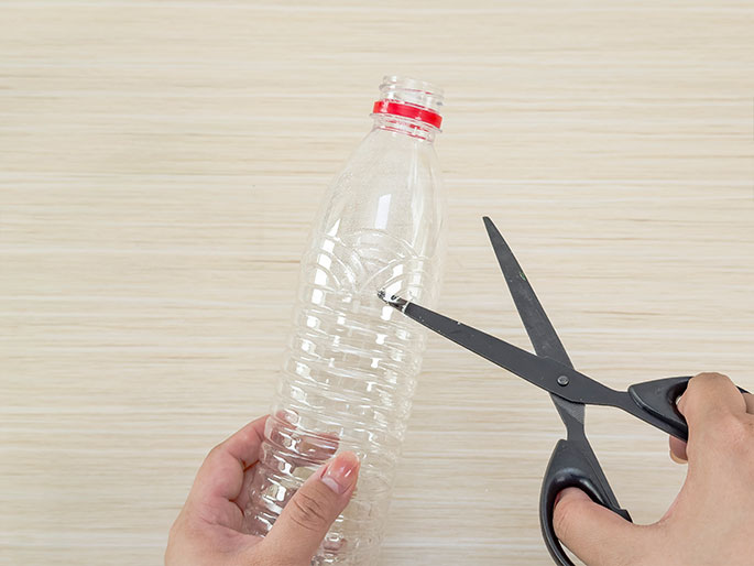 Two hands are shown in the process of cutting a clear plastic bottle with a pair of black scissors. One hand holds the bottle against a light wood background, while the other hand operates the scissors, making a small initial cut in the side of the bottle. The bottle has a red cap and slight textured ridges.