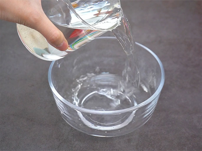 A hand pours water from a clear glass measuring cup into a clear glass bowl. The bowl sits on a dark gray surface. The water flows smoothly from the cup into the bowl. A small section of colorful tape is visible on the measuring cup.