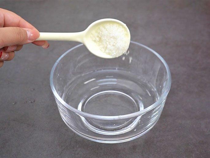 A hand holds a cream-colored plastic spoon filled with white granules, likely salt or sugar, over a clear glass bowl containing water. The spoon is tilted, and some of the granules are falling into the water. The bowl rests on a dark gray surface. The hand has light pink nail polish.