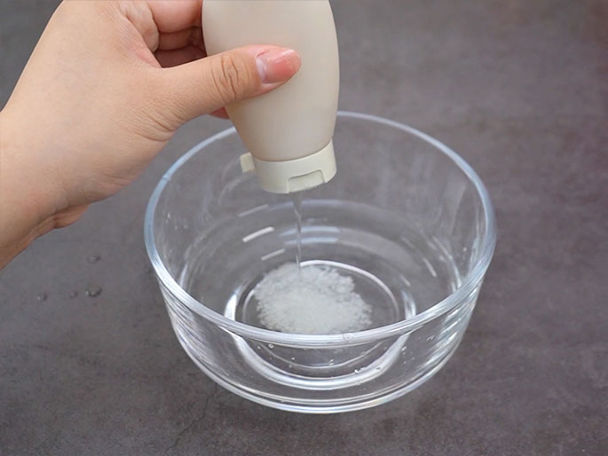 A hand squeezes a small, beige, plastic bottle with a flip-top cap over a clear glass bowl containing water and a dissolved white substance. A thin stream of the bottle's contents is being dispensed into the bowl, adding to the dissolved substance at the bottom. The bowl sits on a dark gray surface.