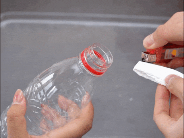 Two hands manipulate items over a gray surface. One hand holds a clear plastic bottle with a red cap. The other hand uses a red and yellow stapler to secure a piece of white material, possibly cloth or paper, around the bottle's opening. The video appears to demonstrate the setup for a science experiment or DIY project, likely involving the creation of bubbles or a chemical reaction within the bottle.