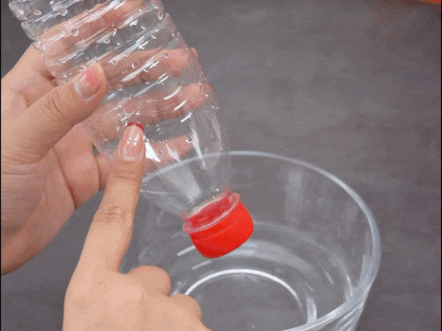 A hand holds a clear plastic bottle with a red cap over a clear bowl filled with water. The hand covers a small hole in the side of the bottle with a finger. The bottle is then lowered, submerging the cap and part of the bottle into the water. The video seems to demonstrate a science experiment or demonstration related to air pressure, water displacement, or a similar scientific principle.