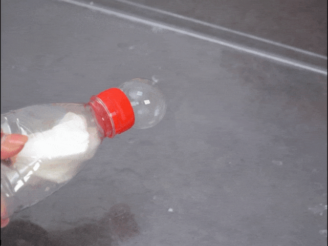 A hand holds a clear plastic bottle with a red cap over a gray surface. The bottle contains a white material, possibly a cloth or paper. A large, clear bubble slowly inflates from the bottle's opening, expanding to nearly the size of the bottle itself. The background is a plain, gray surface, likely a tray or table. This video appears to showcase a science experiment or DIY project involving creating bubbles, possibly using dry ice or a chemical reaction.