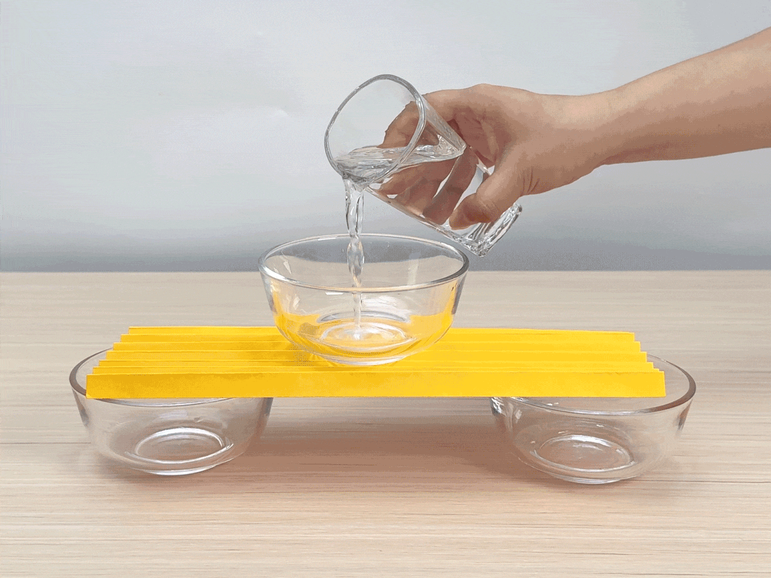 A simple balance scale is constructed using a flat, yellow piece of cardboard or wood as the beam. Two small, clear glass bowls serve as the scale pans, placed at either end of the beam. The beam rests on two more small, empty glass bowls placed upside down. A hand pours water from a clear glass into the bowl on one side of the scale. The added water causes that side of the scale to lower, demonstrating the basic principles of weight and balance. The video depicts a simple physics experiment illustrating how a balance scale works.