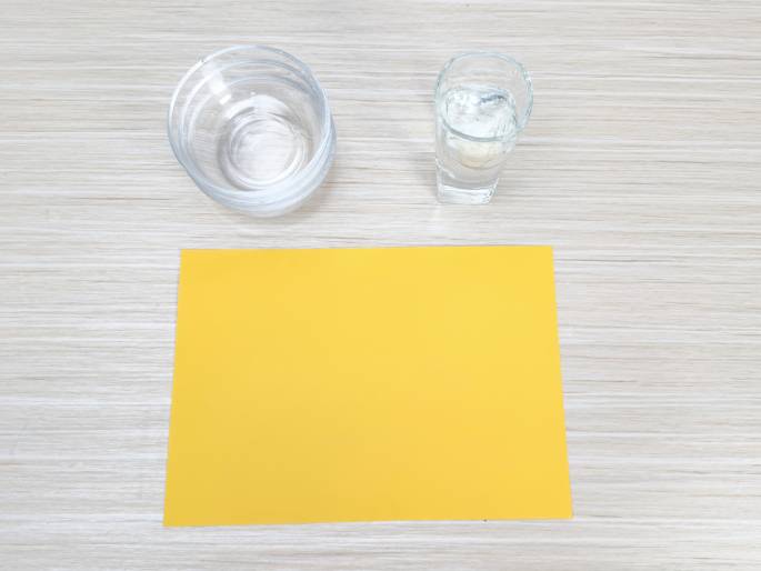 A flatlay image shows a simple arrangement on a light wood-grained table. A small stack of clear glass bowls, a small clear glass filled with water, and a blank sheet of bright yellow paper are positioned in the frame. The scene suggests preparation for a craft or educational activity.