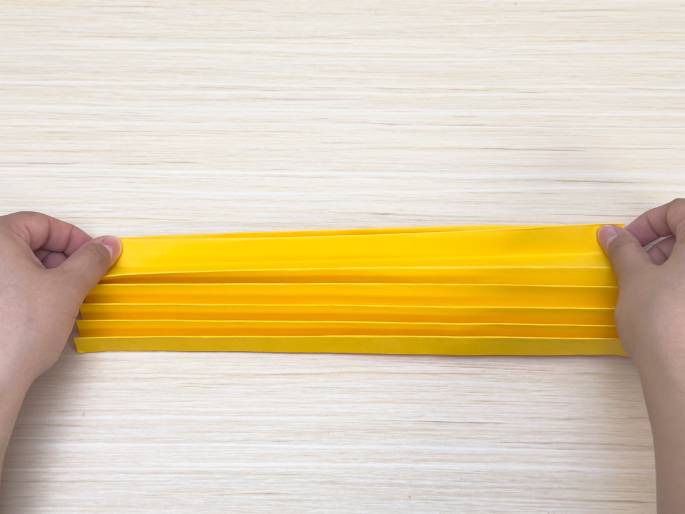 Two hands hold a piece of bright yellow paper that has been folded into a fan or accordion-like shape. The folds are clearly visible, and the hands gently stretch the paper to display the pleats. The paper rests on a light wood-grained surface. This image likely demonstrates a step in a paper craft or origami project.