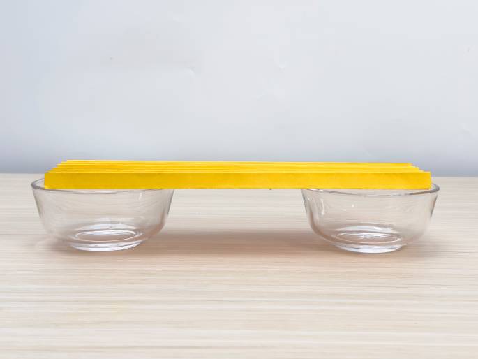 A simple structure is depicted on a light wood-grained table. Two small, clear glass bowls are placed a short distance apart, and a stack of folded yellow paper, resembling a flat, rectangular beam, bridges the gap between them. The bowls appear empty. This image likely represents a basic engineering or physics experiment demonstrating concepts like load-bearing or structural integrity.
