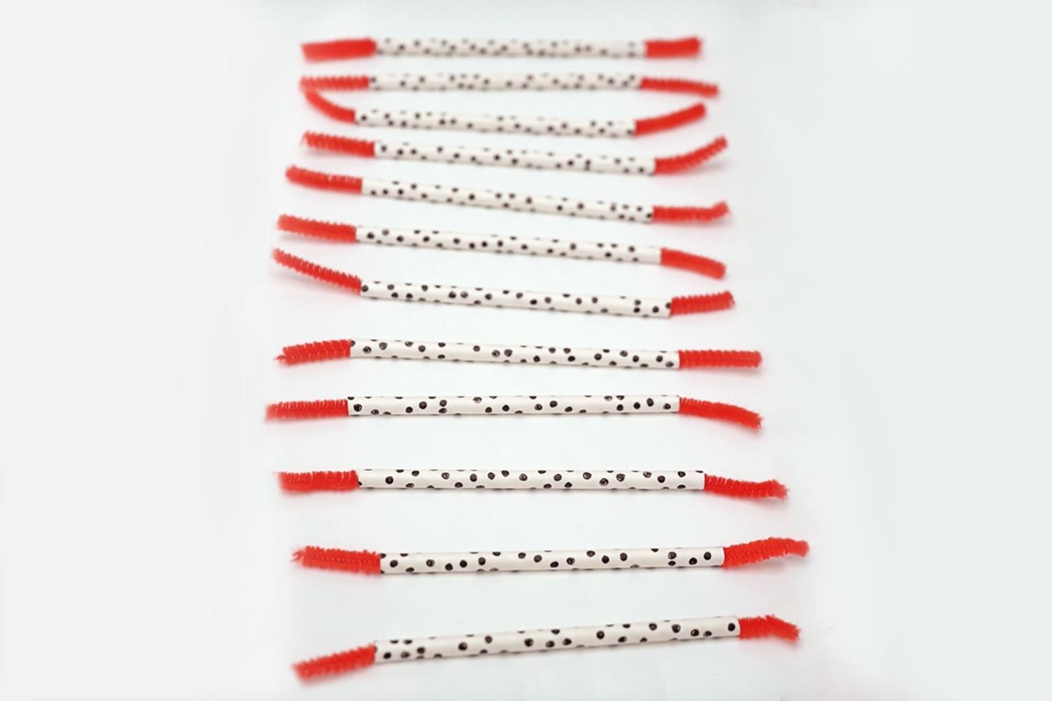 Eleven lengths of white-with-black-spots paper straws are neatly arranged in a row, each with a short length of red pipe cleaner attached to each end on a white surface.