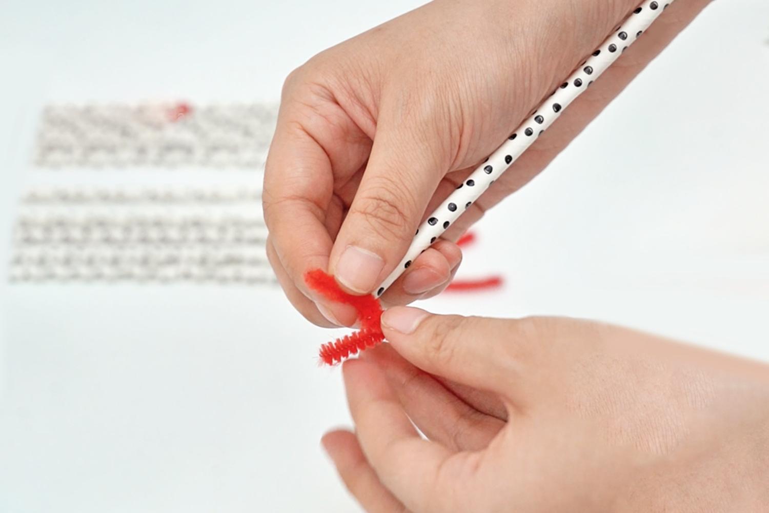 A person's hands carefully attach a short length of red pipe cleaner to one end of a black-polka-dotted paper straw.