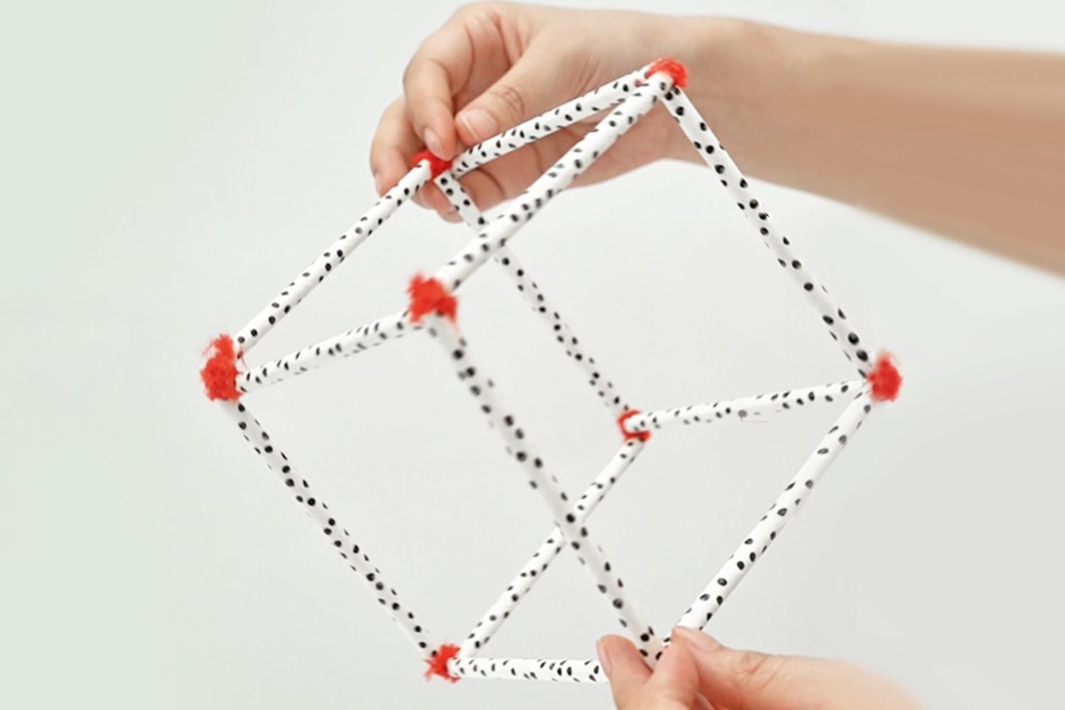 Two hands hold up a cube framework constructed from black-polka-dotted straws joined at the corners with small pieces of red pipe cleaner.