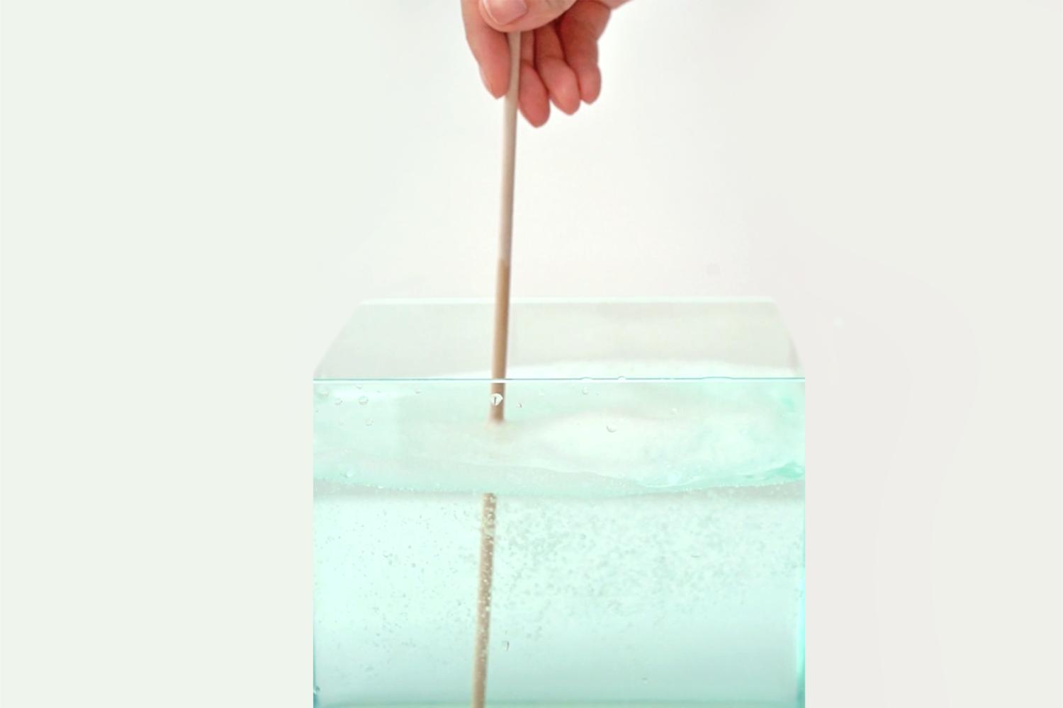 A hand dips a wooden stick into a cube-shaped container of pale green, bubbly liquid against a white background.