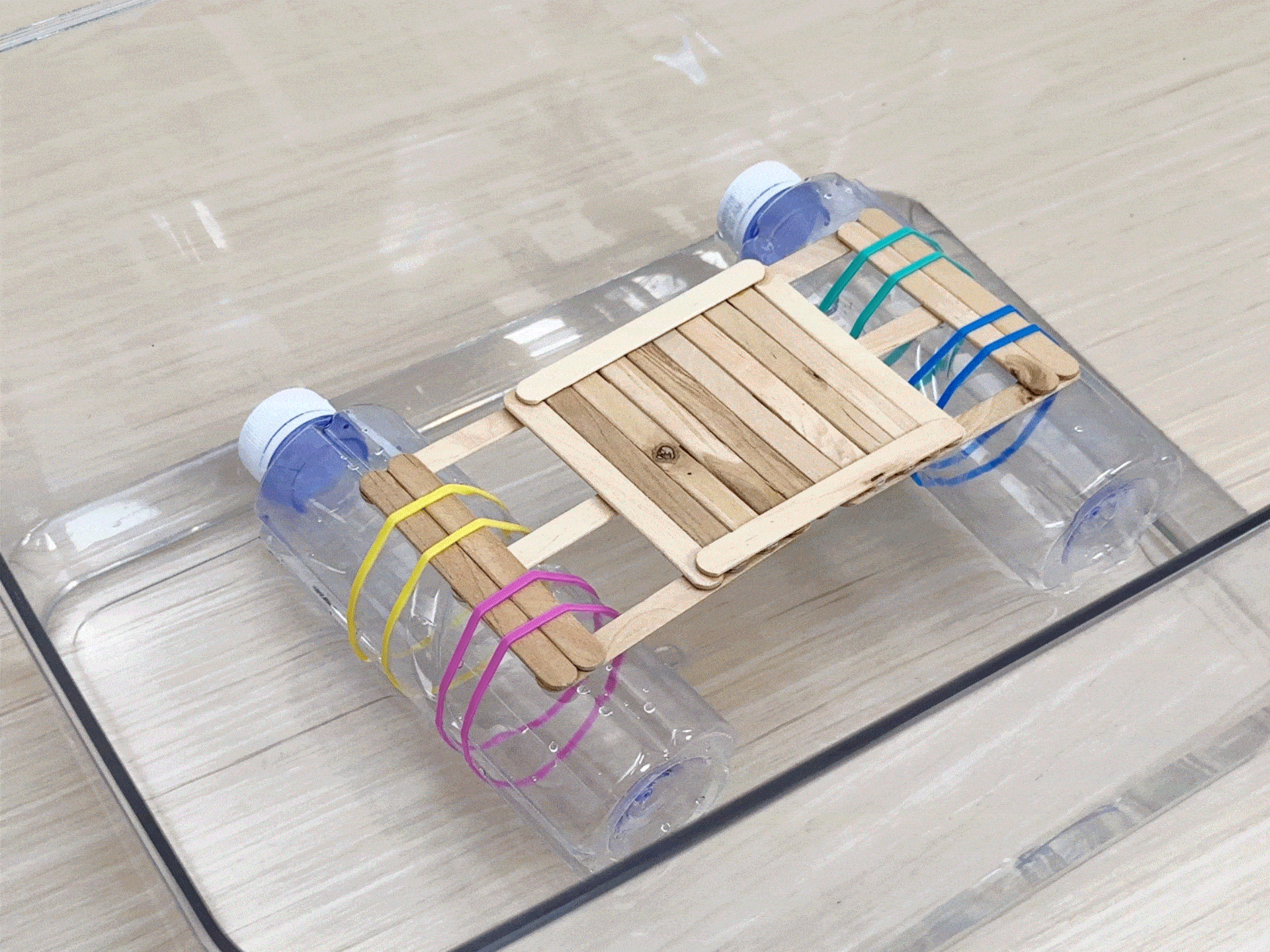 A small raft made of plastic bottles, craft sticks, and rubber bands floats in a clear plastic container filled with water. Rocks are placed on the raft one by one to test its buoyancy.