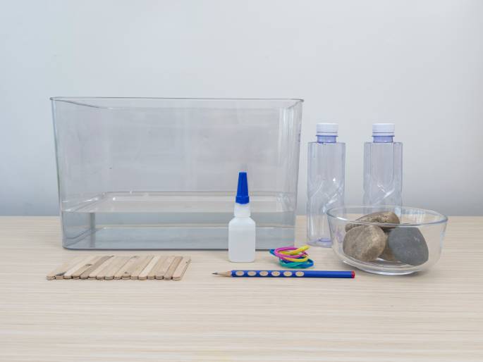 An array of items set up on a light wooden table suggests preparation for a science experiment or craft project. A clear plastic container filled with water sits at the back. In front of it are two empty clear plastic bottles, a small bottle of white glue with a blue cap, a collection of colorful rubber bands, a pencil, a neat stack of small wooden craft sticks, and a clear glass bowl containing several rocks. This arrangement indicates a hands-on activity involving water, buoyancy, or construction.