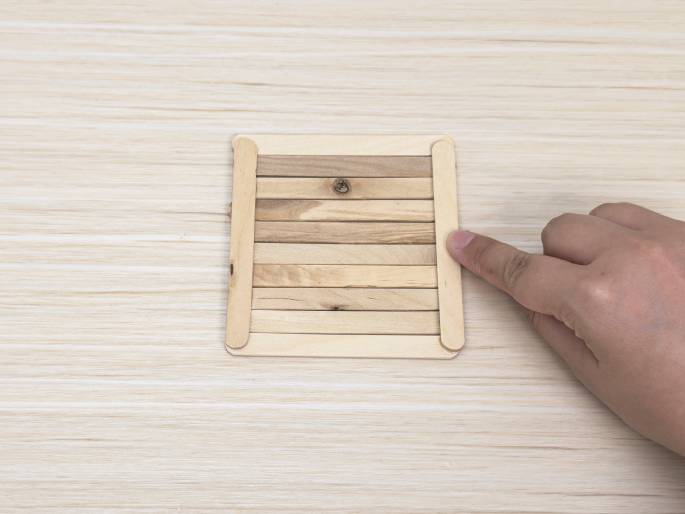 A small raft or platform constructed from wooden craft sticks rests on a light wood grain surface. The sticks are laid side-by-side and glued together, forming a rectangular shape. Two additional sticks are glued perpendicularly along the longer sides, acting as a frame or reinforcement. A finger points towards the lower right corner of the raft. This image likely depicts a stage in a crafting project.