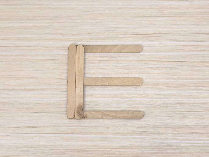 The capital letter E is formed on a light-colored wooden background using wooden craft sticks. Four sticks are arranged to create the letters shape. Two slightly overlapping sticks form the vertical left side, and three shorter sticks create the horizontal top, middle, and bottom bars.