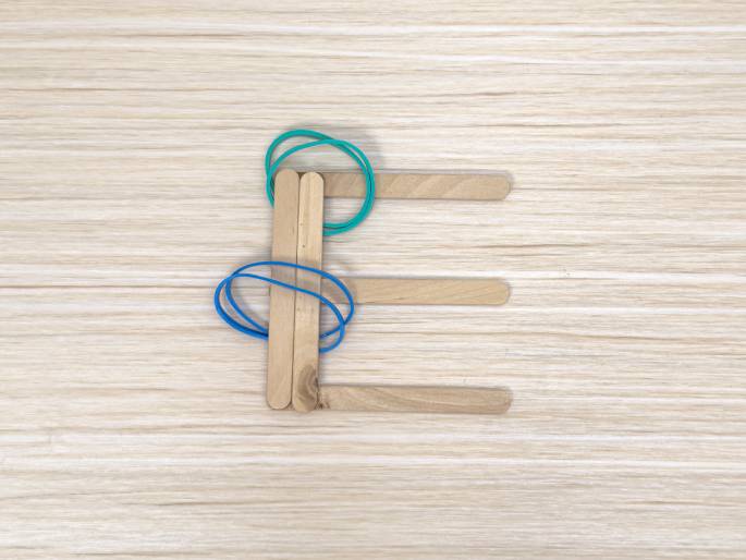 The capital letter E is constructed from wooden craft sticks on a light wood background. Two craft sticks, slightly overlapping, form the vertical part of the E, and are bound together with a blue rubber band. A teal or green rubber band is looped around the top of these two sticks. Three additional sticks are positioned horizontally to create the top, middle, and bottom bars of the E.
