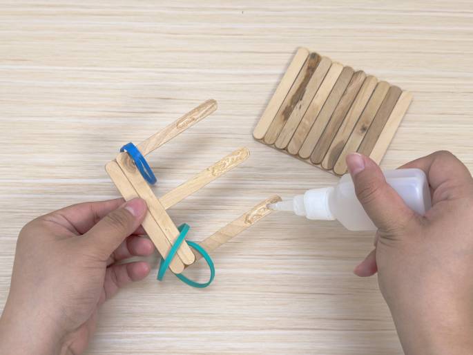 Two hands work on a craft project on a light wooden surface. One hand holds a structure made of craft sticks, secured with blue and teal rubber bands. The other hand applies glue from a small white bottle to the top of one of the sticks. A separate, completed section of the project, a square made of glued craft sticks, lies on the table to the right. This image captures the process of assembling a craft using wooden sticks, glue, and rubber bands.