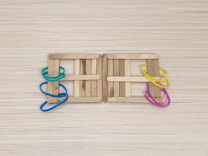 Two rectangular frames made of wooden craft sticks sit side-by-side on a light wood grain surface. They appear to be connected, or hinged, at the center. Colorful rubber bands are wrapped around the craft sticks on the outer edges of each frame. A blue and green rubber band are on the left frame, and yellow and pink rubber bands are on the right frame. This arrangement suggests a homemade toy or a craft project in progress.