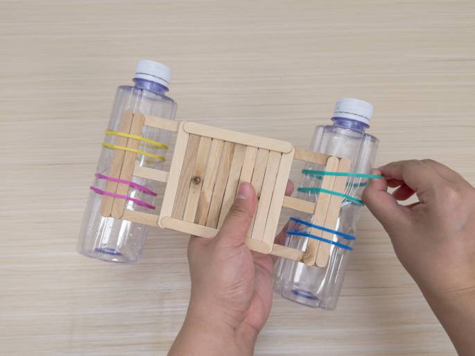 Hands hold a simple raft or boat constructed from recycled materials against a light wood background. The boat's base is a platform made of craft sticks glued together. Two clear plastic bottles are attached to the underside of the platform, one on each end. Craft sticks are fastened to the bottles with colorful rubber bands—pink and yellow on one bottle, and teal and blue on the other. One hand holds the boat while the other adjusts a rubber band. This image likely depicts a children's craft project or a simple science experiment demonstrating buoyancy.