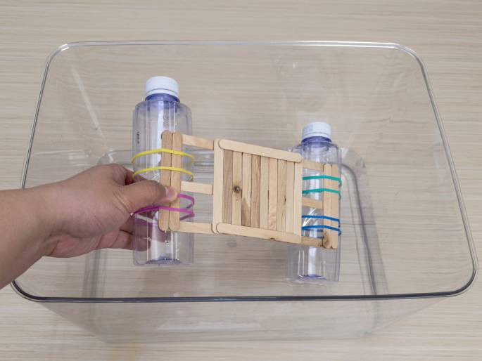 A hand holds a small raft made of recycled materials over a clear plastic container. The raft is constructed with a platform of glued craft sticks and two clear plastic bottles attached underneath, one at each end. Colorful rubber bands—pink and yellow on one bottle, and teal and blue on the other—secure craft sticks to the bottles. The container sits on a light wood surface, and it appears to be empty, ready for water or another testing medium. This image likely depicts a science experiment demonstrating buoyancy or a children's craft project.