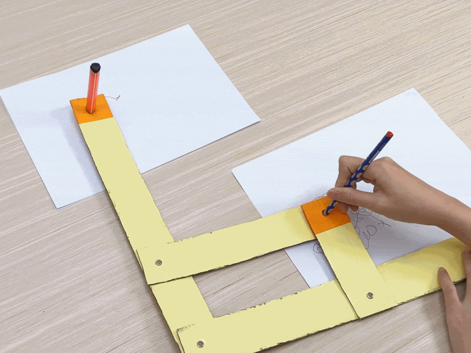 A child uses a simple drawing machine made of yellow painted cardboard strips and fasteners. Markers and pencils are placed through holes in the cardboard and used to draw shapes on separate sheets of paper.