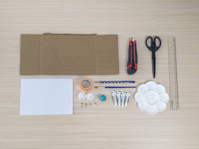 An assortment of craft supplies lies neatly arranged on a light wood grain surface. The items include a piece of corrugated cardboard, a blank white sheet of paper, a roll of orange masking tape, two paintbrushes, a pencil, small tubes of glue or paint, a flower-shaped paint palette, a utility knife, scissors, two white plastic lids, thumbtacks, a small ball of blue modeling clay, and a clear plastic ruler. These materials appear ready for a crafting project.