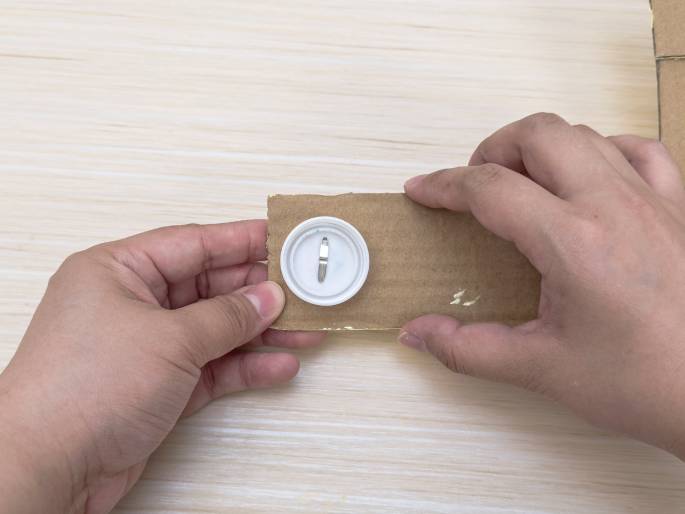 Two hands hold a small rectangular piece of plain brown cardboard on a light wooden surface. A white plastic bottle cap is attached to the cardboard with a metal brad or fastener. The prongs of the fastener are visible on the top of the bottle cap. This image seems to depict a stage in a crafting process.