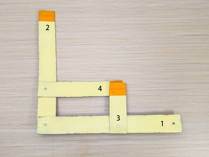 Four pale yellow cardboard strips, numbered 1 through 4, are arranged on a light wood grain surface to form an incomplete square. Strips 1 and 2 form an L shape, while strips 3 and 4 form a smaller L connected to the first. The ends of strips 2 and 4 are covered with bright orange material. Small metal brads or fasteners are visible at the points where the strips overlap. This appears to be another stage in the construction of a cardboard object.