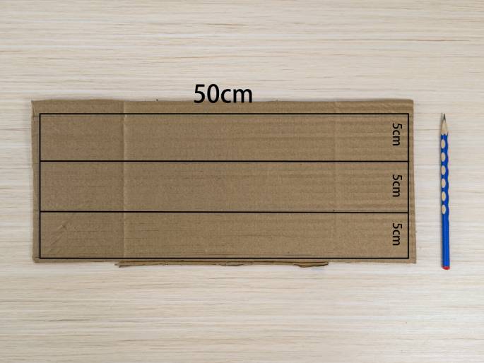 A rectangular piece of cardboard rests on a light wood grain table. Three horizontal lines, spaced 5cm apart, have been drawn across the cardboard, dividing it into four equal sections. The top edge of the cardboard is labeled 50cm, indicating its length. A blue pencil lies next to the cardboard, suggesting it was used for marking. This image likely depicts the initial steps in a crafting project.