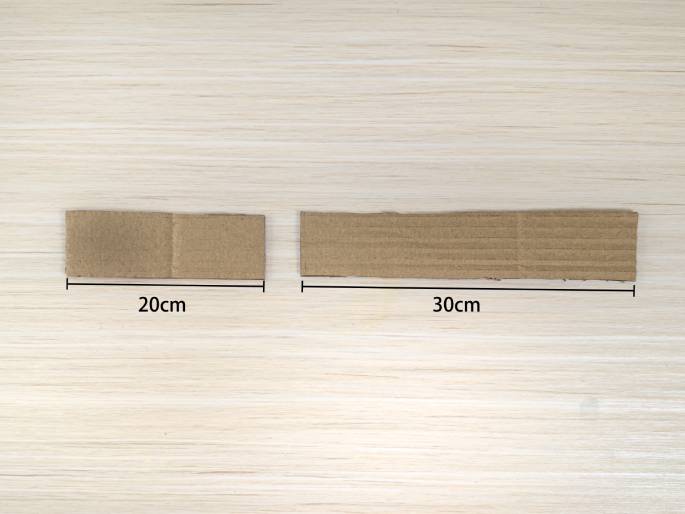 Two pieces of cardboard lie on a light-colored wooden surface. The smaller piece, labeled 20cm, is positioned to the left. The larger piece, labeled 30cm, is to the right. These pieces appear to have been cut for a crafting project.