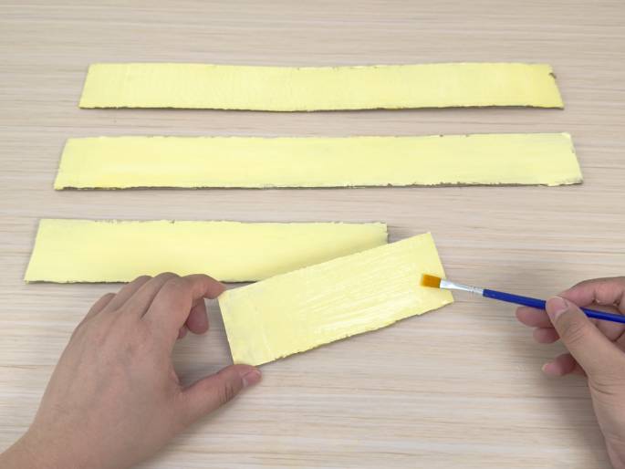 Three strips of cardboard, painted pale yellow, are arranged on a light wood grain table. Two of the strips are fully painted and lie flat, while a third strip is being painted by a hand holding a small blue paintbrush. The hand holds the strip at an angle as yellow paint is applied.