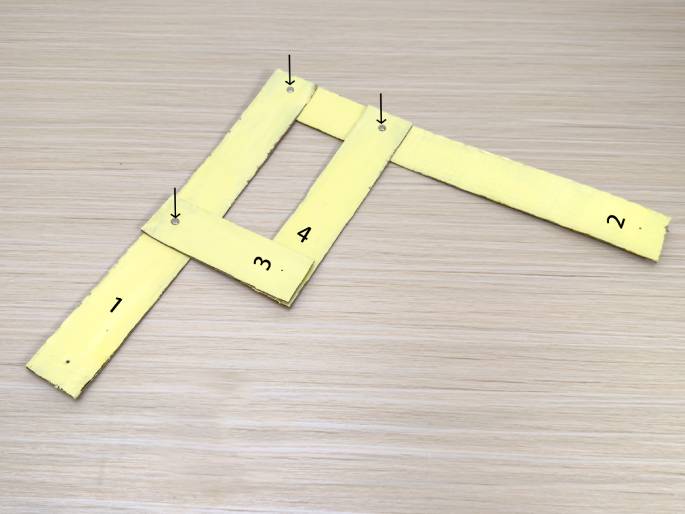 Four pale yellow cardboard strips are arranged on a light wood grain surface, connected by small fasteners at their overlapping points. The strips are numbered 1 through 4. Strips 3 and 4 form a rectangular frame, with strip 1 extending downwards to the left and strip 2 extending diagonally upwards to the right. Arrows point towards the fasteners holding the strips together. This image appears to show a partially assembled cardboard construction.