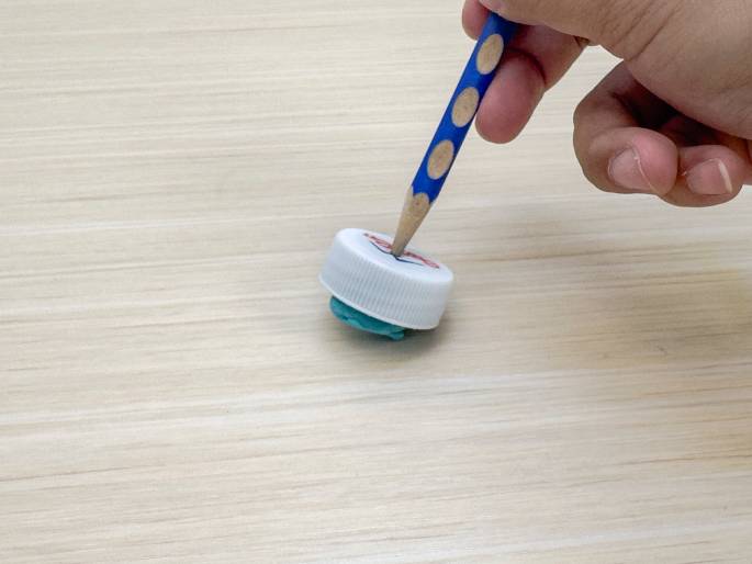 A white plastic bottle cap, with teal-colored modeling clay or putty around its base, sits on a light wood grain surface. A hand holds a pencil, which is piercing the center of the bottle cap. This setup appears to be a homemade toy or a simple craft project.