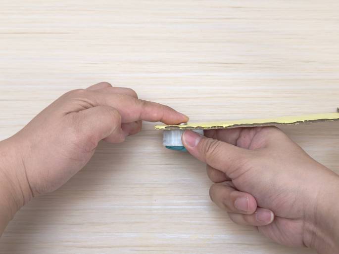 Two hands manipulate a piece of pale yellow cardboard and a white plastic bottle cap with teal putty on a light wood grain surface. The bottle cap, with the putty acting as an adhesive, is being attached to the underside of the cardboard. A small, metallic brad or fastener is visible, securing the bottle cap to the cardboard. One hand holds the cardboard strip while the other hand positions the bottle cap and presses it against the cardboard.