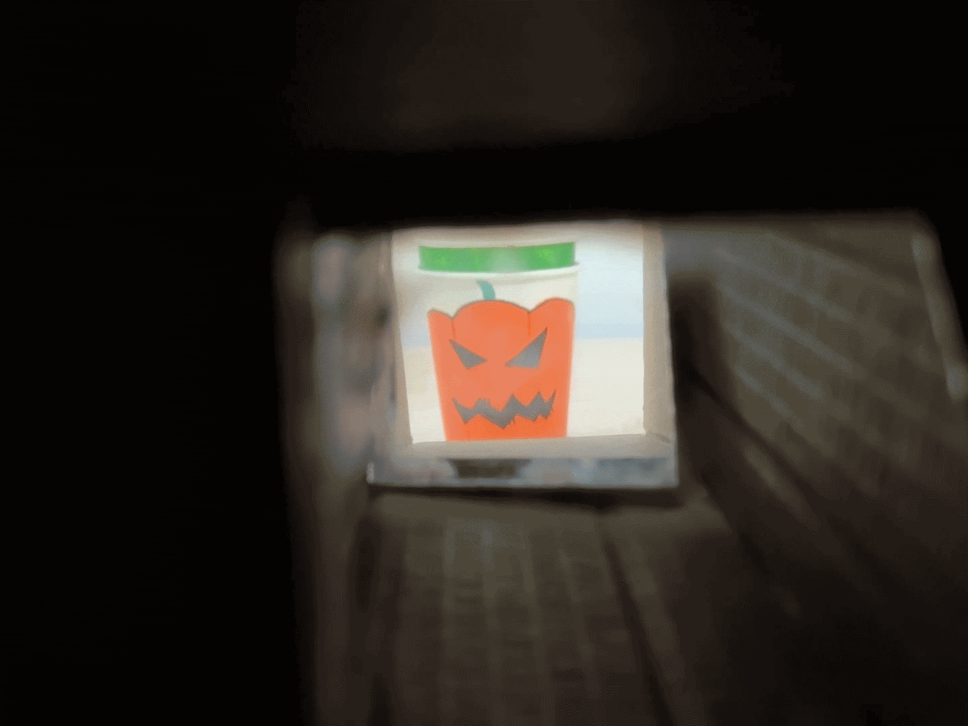 The camera looks through a cardboard tube at an orange paper cup with a jack-o'-lantern face. The camera pulls back to reveal the cup on a wooden table.