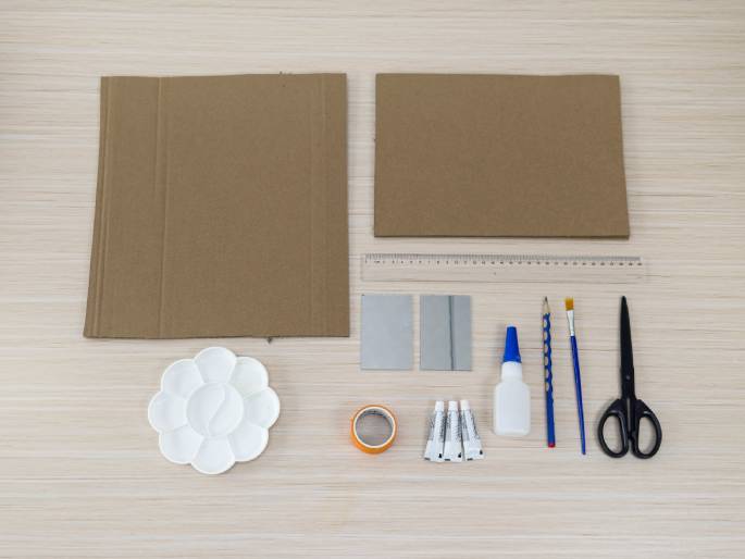 An assortment of craft supplies lies neatly arranged on a light wood-grain table. Two pieces of brown cardboard are at the top, followed by a ruler, two small mirrored tiles, a white flower-shaped paint palette, a roll of orange tape, three small tubes of adhesive, a bottle of white glue, a pencil, a paintbrush, and a pair of scissors. The items are laid out in a flat lay style, ready for use in a craft project.