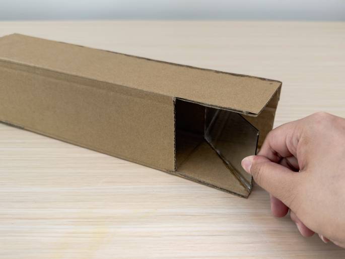 A long rectangular box made of brown cardboard sits on a light wood-grain table. A hand is inserting a small mirror into a cut-out section near one end of the box. The mirror is angled within the box. The box appears to be a homemade project, possibly a periscope or a simple optical device.