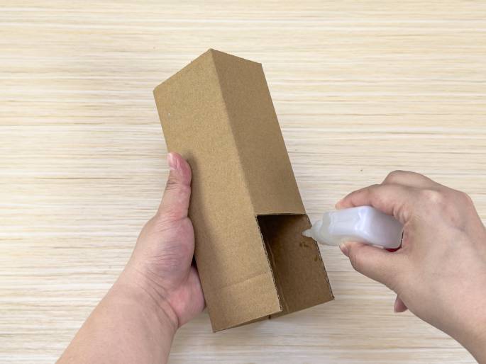 Two hands are working with a rectangular cardboard box on a light wood-grain table. One hand holds the box, which has an open top and a square cut-out section near the bottom. The other hand applies glue from a small white bottle into the interior of the cut-out section. The box appears to be a homemade project in progress.