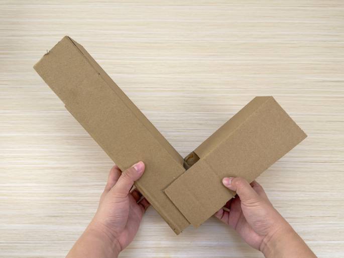 Two hands hold two rectangular cardboard boxes positioned to form a V shape on a light wood-grain table. The longer box lies diagonally, while the shorter box is attached to it at a right angle. The hands appear to be assembling or connecting the two boxes, suggesting a DIY project.