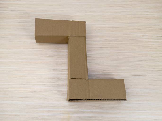 Two rectangular cardboard boxes are joined together to form the shape of the letter L on a light wood-grain surface. The boxes are made of plain brown cardboard, and the joint where they connect appears glued or fastened. This suggests a DIY project or a repurposed packaging material.
