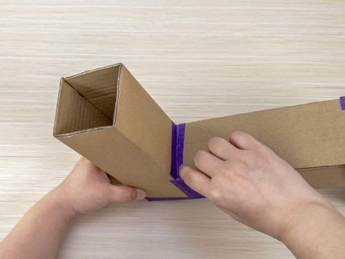 Two hands are assembling a structure made of brown cardboard boxes on a light wood-grain table. The boxes are arranged in an L shape. Purple tape is being used to secure the joint where the two boxes meet. The open end of one box reveals a square opening, while the other box lies flat. This image depicts a DIY project in progress.
