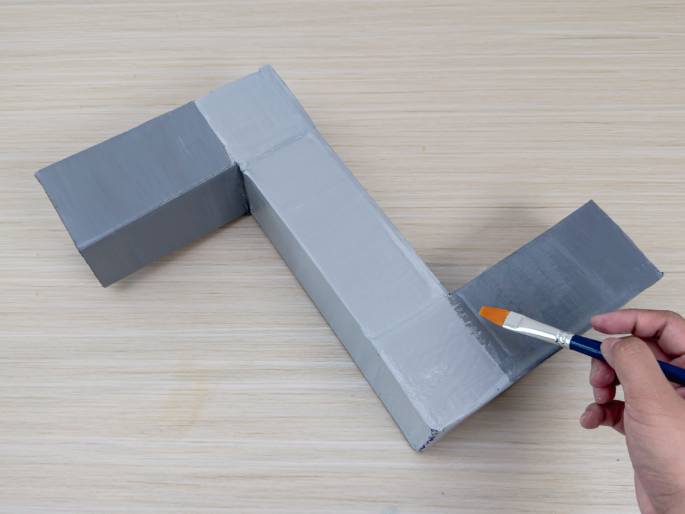 An L-shaped object constructed from cardboard boxes is being painted gray on a light wood-grain table. A hand holds a paintbrush with an orange bristle tip and a blue handle, applying gray paint to the cardboard. Some sections appear already painted, while others are in progress, indicating the project is not yet finished.