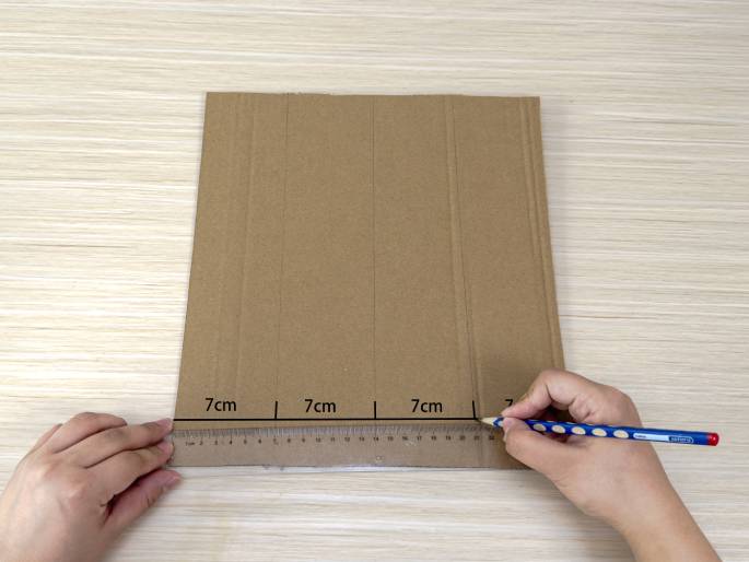 Two hands are shown measuring and marking a piece of cardboard. One hand holds a clear plastic ruler against the bottom edge of the cardboard, while the other hand uses a pencil to draw a line along the ruler. The cardboard rests on a light wood-grain table. Three 7cm markings are visible along the bottom edge, indicating measurements for cutting or folding.